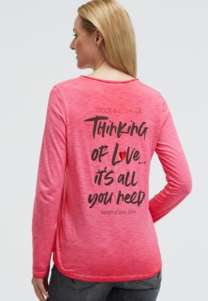 Langärmliges, pink meliertes Shirt mit abgerundetem Saum. Auf der Rückseite steht in schwarzer Schrift: "Thinking of Love... it's all you need," mit einem Herzakzent.