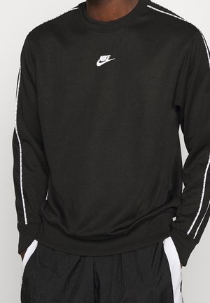 Sort Nike langærmet skjorte med hvidt logo på brystet og hvidstribet bånd langs ærmerne, båret med sorte bukser med hvide sidestriber.