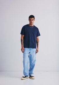 Un tricou bleumarin cu mânecă scurtă combinat cu blugi decolorati, cu croială relaxată și teniși negri cu talpă crem, stând pe un fundal alb.