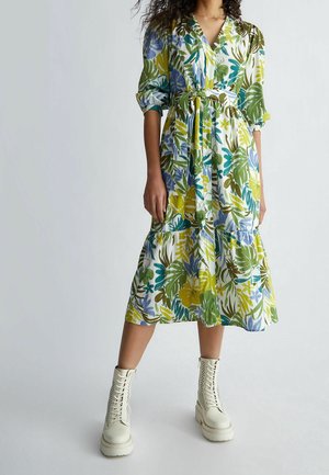 Femme portant une robe fleurie mi-longue avec des feuilles vertes, bleues et jaunes, associée à des bottes blanches à plateforme lacées.