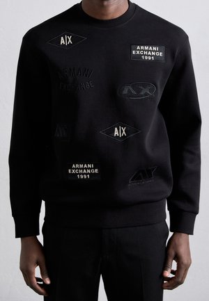 Zwarte trui gedragen door persoon, met meerdere geborduurde Armani Exchange-logo's en tekstpatches in wit en zwart.