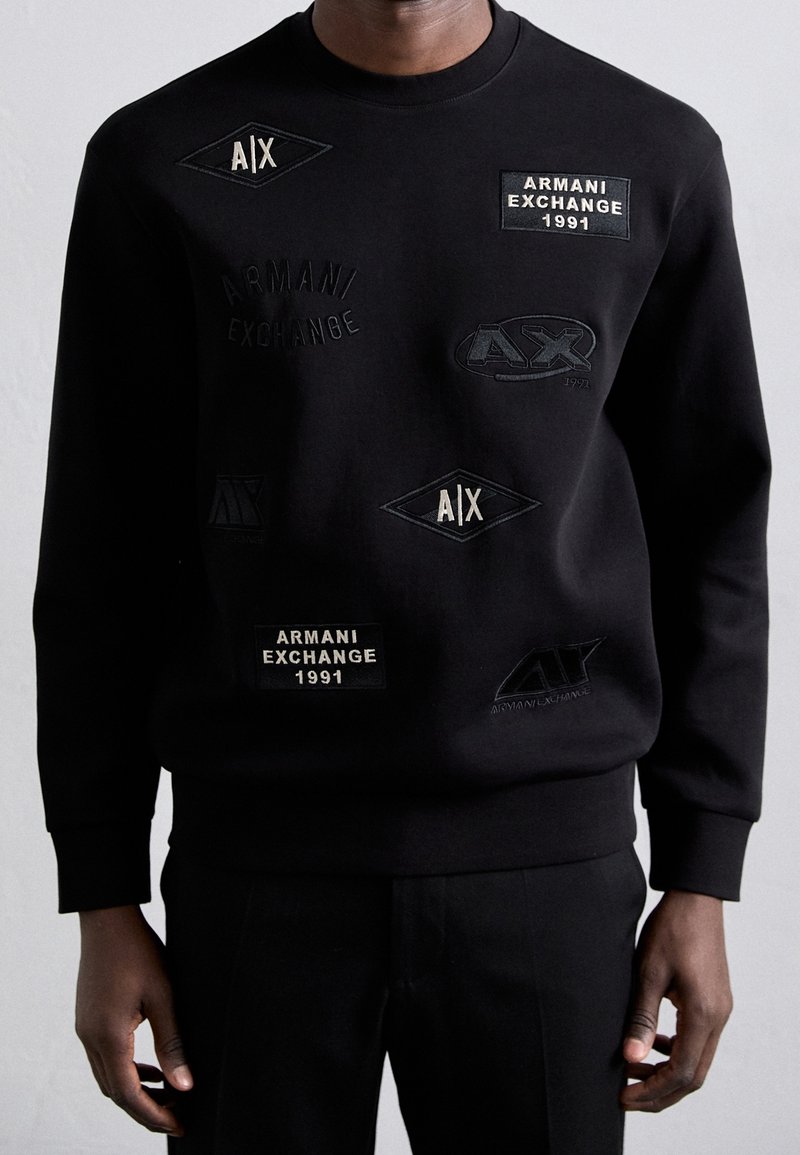 Sweat-shirt noir porté par une personne, avec plusieurs logos brodés Armani Exchange et des patchs de texte en blanc et noir.