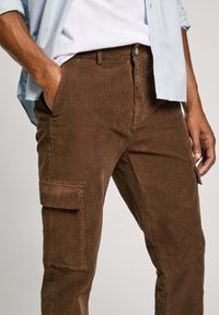 Pantalones cargo de pana marrón con bolsillos laterales, cierre de botón y textura sutil. Combinados con una camisa azul clara.