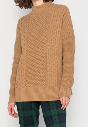 Beżowy sweter z dzianiny z prążkowanymi mankietami i dołem, teksturowanym wzorem w splot warkoczowy oraz wysokim kołnierzem. Noszony na ciemnych spodni z kratkę.