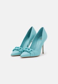 Turquoise high heels made of smooth leather, featuring a pointed toe and a decorative knotted accent across the front. Slim stiletto heels.