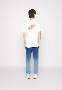 Bílý tričko s růžovým a černým logem Nike na zadní straně, ve spojení s modrými gradientovými tepláky a bílými teniskami s barevnými akcenty.