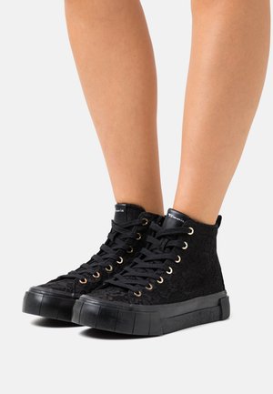 Voeten met zwarte hoge sneakers met veters, dikke rubberen zolen en zwarte veterdetails, afgebeeld tegen een effen lichte achtergrond.