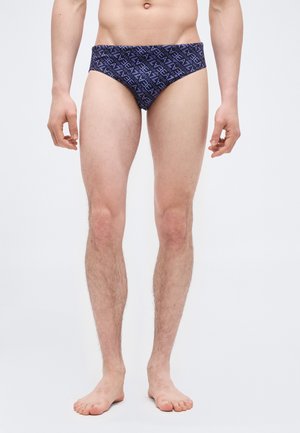 Άνδρας όρθιος ξυπόλητος φορώντας σκούρα μπλε εσώρουχα με επαναλαμβανόμενο λευκό κείμενο σε απλό φωτεινό φόντο.