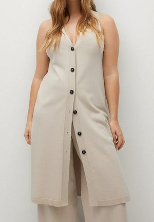 Beżowa, bezrękawnikowa długa kamizelka zapinana na guziki, noszona na spodniach o szerokich nogawkach w tym samym kolorze, prezentowana na osobie stojącej z rozpuszczonymi włosami.