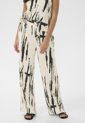 Vrouw draagt crèmekleurige broek met wijde pijpen en bijpassend top met zwart abstract penseelstreekpatroon en zwarte sandalen.