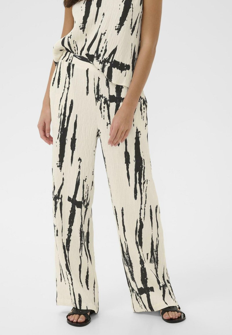 Femme portant un pantalon large crème et un haut assorti avec un motif noir abstrait en coups de pinceau, ainsi que des sandales noires.