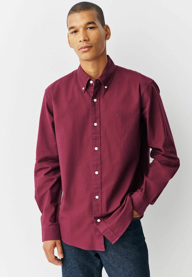 Cămașă burgundy cu butoni, cu mâneci lungi, butoane albe și un mic logo pe pieptul stâng. Fabricată dintr-un material neted, croială ajustată.