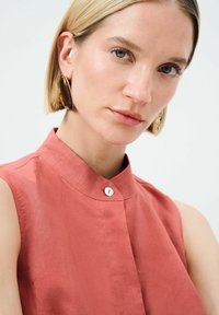 Rosa ärmelloses Shirt mit hohem Kragen, das ein kleines Knopfdetail am Hals aufweist und aus einem weichen, strukturierten Stoff besteht.