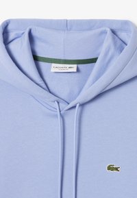 Sudadera azul clara hecha de una tela suave, con un bolsillo canguro en la parte frontal y un pequeño logo de cocodrilo verde en el pecho. Incluye cordones.