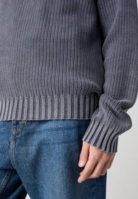 Nahaufnahme einer Person, die einen grauen Strickpullover und blaue Jeans trägt, mit der rechten Hand entspannt an der Seite.