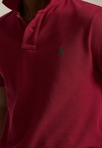 Homme portant un polo rouge foncé avec un logo vert foncé brodé sur le côté gauche de la poitrine et le col partiellement relevé.