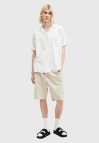 Hvid kortærmet skjorte med tekstureret mønster, cremefarvede shorts, hvide sokker og sorte sandaler med dobbeltstropper. Ensfarvet baggrund.