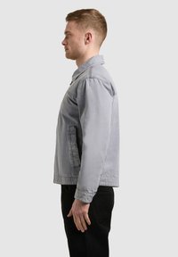 Homme debout de profil portant une veste gris clair et un pantalon noir sur un fond clair uni.