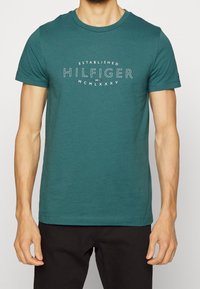 Teal puuvillane T-särk lühikeste varrukatega, ümmargune kaelus ja ees tekstigraafika: "ASUTATUD HILFIGER MCMLXXXV" valgete ja punaste aktsentidega.