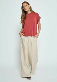 Raudona trumpomis rankovėmis marškinėliai su laisvu siluetu, derinami su kreminės spalvos plačiomis kelnėmis, turinčiomis priekinės klostes ir kišenes. Dėvimos juodos šlepetės.