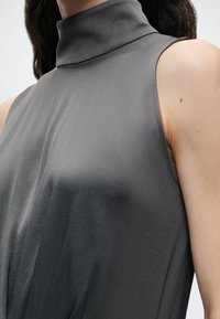 Haut sans manches gris foncé avec col haut porté par une personne aux longs cheveux foncés, montrant les épaules et le haut de la poitrine.