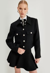 Chaqueta de tweed negra con cuello, dos bolsillos delanteros y botones decorativos de perlas, combinada con una falda negra acampanada. Acabado texturizado.