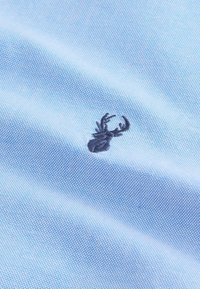 Tissu bleu clair avec un motif texturé à pois, mettant en avant un design de crabe brodé en bleu foncé. Le tissu semble lisse et respirant.
