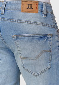 Jean en denim bleu clair avec une texture légèrement usée, deux poches arrière et une étiquette rectangulaire en tan avec le logo.