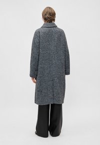 Pelēka zīda šuvītēm mētelis ar izgriezumu un brīvu piegriezumu, kas piedāvā garas piedurknes un fakturētu audumu, valkāts virs melnām platām biksēm.