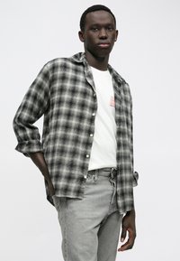 Νέος άνδρας με σκούρο δέρμα, φορώντας γκρι καρό πουκάμισο πάνω από λευκό t-shirt και γκρι τζιν, στέκεται με το ένα χέρι στην τσέπη του, μπροστά σε λευκό φόντο.