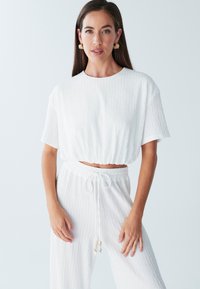 Biały, teksturowany krótki top z krótkimi rękawami, zestawiony z białymi, luźnymi spodniami na sznurek. Charakterystyczne koraliki na sznurku.