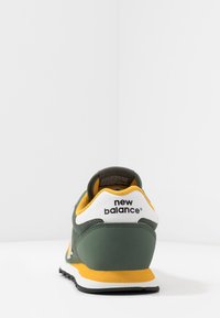 Scarpa da atletica verde e gialla realizzata in materiali mesh e suede, con una linguetta posteriore bianca e una suola in gomma nera. La vista posteriore mostra il marchio.
