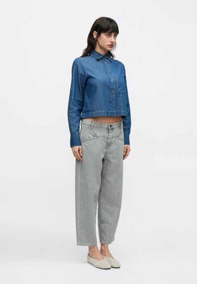 Chemise en jean raccourcie bleu moyen avec boutons, associée à un jean ample taille haute gris clair et des chaussures beige à enfiler, posant sur un fond uni.