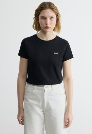 Γυναίκα με κοντά ξανθά μαλλιά, φορώντας μαύρο μπλουζάκι BOSS και ψηλόμεσο λευκό παντελόνι, στέκεται μπροστά σε απλό φωτεινό φόντο.