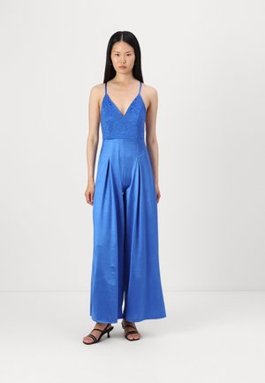 Combinaison bleue avec un corsage en dentelle, décolleté en V et design à jambes larges. La texture soyeuse et les détails plissés à l'avant complètent le style moderne. Sandales noires.