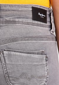Los jeans de mezclilla gris presentan un bolsillo trasero con detalles de costura blanca y una etiqueta de cuero negro que dice "Pepe Jeans London."