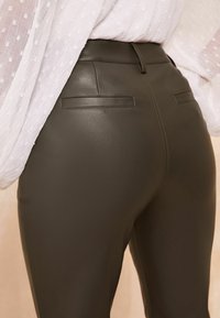 Pantaloni din piele ecologică de culoare verde închis, cu talie înaltă și ajustați, prevăzuți cu două buzunare pe spate și detalii de cusătură vizibile. Textură netedă cu un ușor luciu.