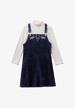 Marineblauwe corduroy overall jurk met verstelbare bandjes en bloemenborduursel, gecombineerd met een geribbelde witte coltrui met gegolfde manchetten.