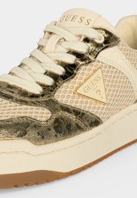 Gros plan d'une basket Guess beige et dorée avec tissu en mesh, lacets blancs, triangle du logo Guess bordé d'or et semelle texturée.