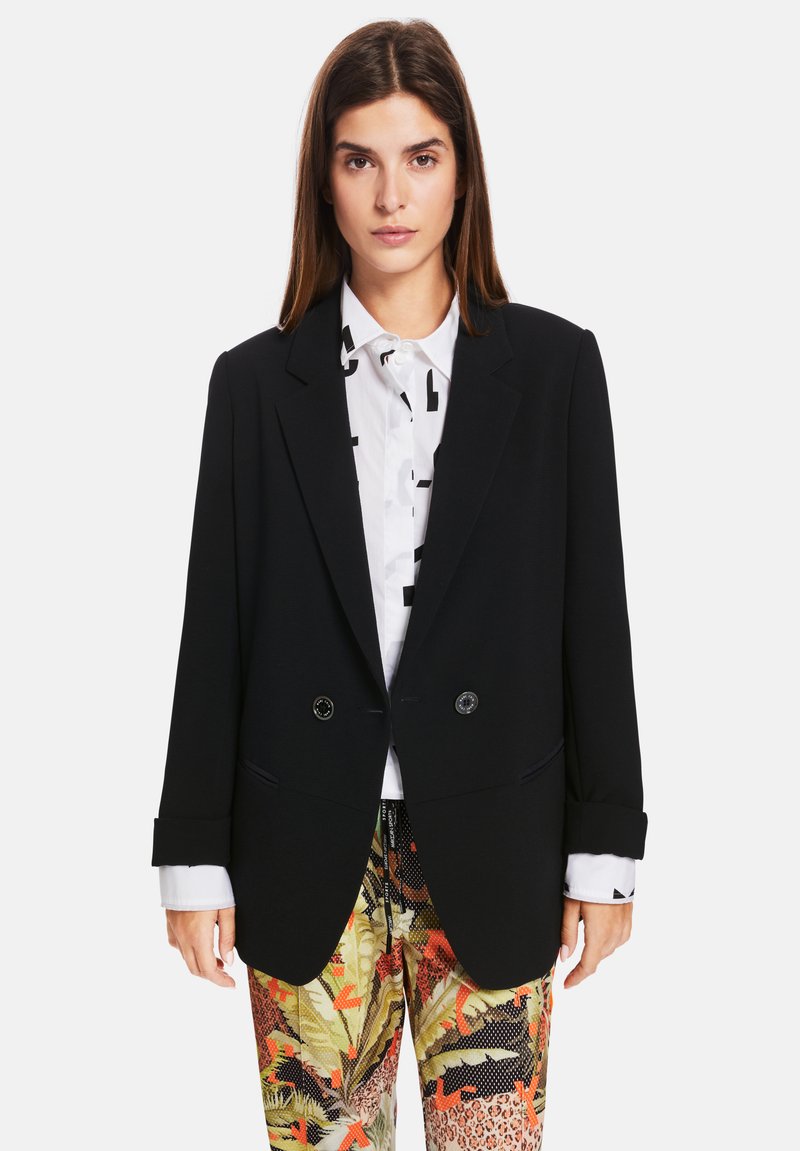 Black double-breasted blazer with notch lapels and button accents, paired with a white shirt featuring abstract black patterns, and colorful patterned pants.