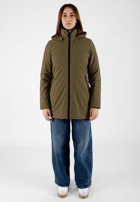 Giacca impermeabile verde oliva con colletto alto e cappuccio, dotata di una cerniera frontale, dettagli neri e due tasche laterali. Abbinata a jeans di denim larghi.