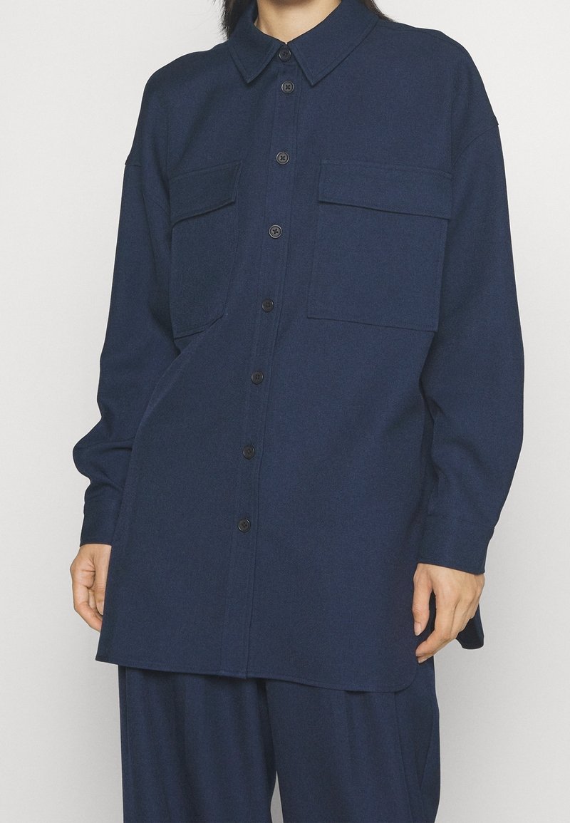 Marinblå knapp-up skjorta i texturerad fabric, med två bröstfickor, krage och en avslappnad passform med böjd nederkant.