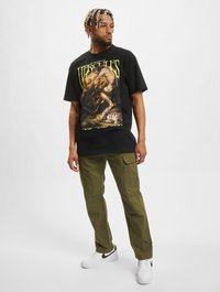 Svart grafisk t-skjorte med Hercules-design og grønn tekst. Kombinert med olivenfargede cargobukser og hvite joggesko. Modell i stående posisjon.