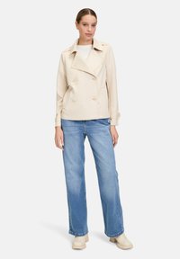 Beżowa podwójnie zapięta kurtka z dużymi guzikami, kieszeniami po bokach i naramiennikami, w zestawieniu z jasnoniebieskimi spodniami z szerokimi nogawkami i beżowymi klapkami.