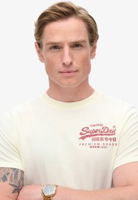 Creme kortærmet t-shirt med et rødt trykt logo, rund halsudskæring og blød stofstruktur. Båret af en person med et armbåndsur.