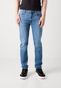 Vīrietis ar gaiši ziliem džinsiem, melnu T-kreklu un melnām kedām ar baltām zolēm, stāv uz vienkārša gaiša fona.