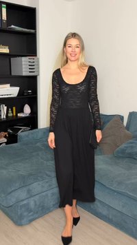 Γυναίκα με μαύρο φόρεμα μακρυμάνικο από δαντέλα και μαύρες γόβες, που στέκεται σε ένα σαλόνι κοντά σε ένα μπλε καναπέ και μια μαύρη βιβλιοθήκη.