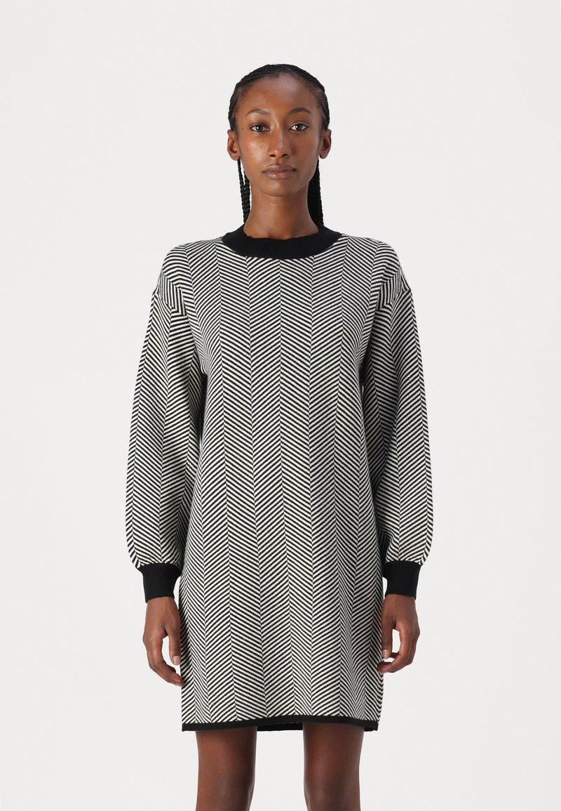 Vestido com padrão zigzag em preto e branco, com gola e punhos ribados em preto, apresentando um corte solto e comprimento até a metade da coxa. Textura suave.