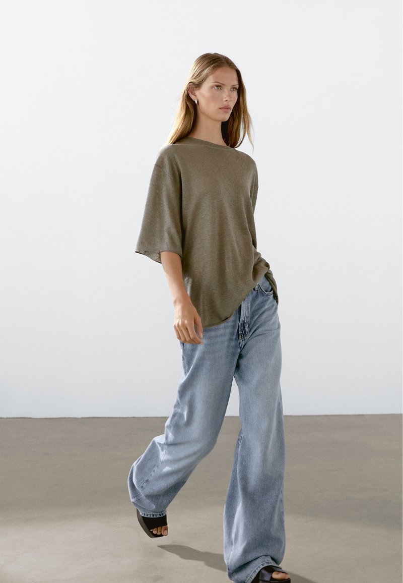 Olivgrön oversized tröja med korta ärmar, kombinerad med ljusblå vida jeans och svarta platåsandaler. Enkelt, texturerat tyg.