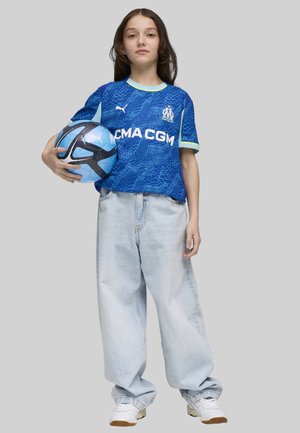 Blå fotbollsjacka med prickigt mönster, ljusgröna accenter och vit logotyp, tillsammans med en oversized ljusblå jeans och vita sneakers, håller en fotboll.